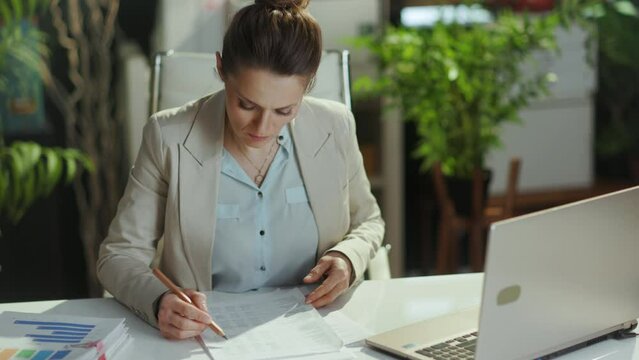 modern business woman in a light business suit in modern green office with documents and laptop.