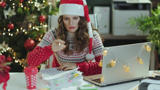 Christmas time. stressed elegant middle aged business woman in santa hat and red Christmas sweater with documents and laptop working in modern green office with Christmas tree.