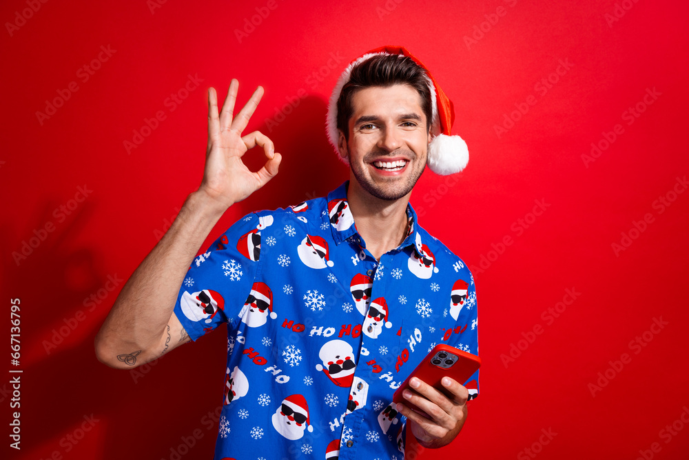 Sticker portrait of satisfied optimistic person dressed santa hat hold smartphone showing okey good job isol