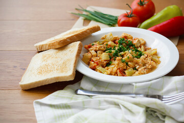 Vegetable dish called lecho containing peppers, tomatoes, onion, eggs and tofu served on white plate with chopped chives and toasted bread. Delicious lecho and its ingredients on wooden table