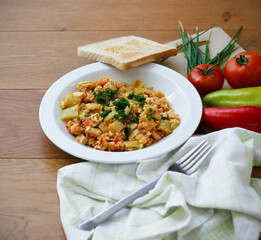 Vegetable dish called lecho containing peppers, tomatoes, onion, eggs and tofu served on white plate with chopped chives and toasted bread. Delicious lecho and its ingredients on wooden table