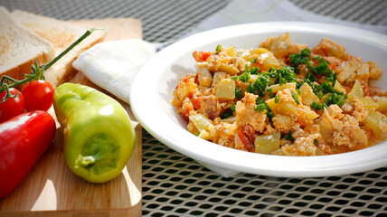 Vegetable dish called lecho containing peppers, tomatoes, onion, eggs and tofu served on white plate with chopped chives and toasted bread. Delicious lecho and its ingredients on garden table.