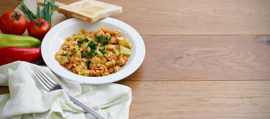 Vegetable dish called lecho containing peppers, tomatoes, onion, eggs and tofu served on white plate with chopped chives and toasted bread. Delicious lecho and its ingredients on wooden table