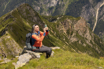 Naklejka na ściany i meble Gray-haired man with backpack sitting on rocky ledge and taking pictures of summer mountain landscape, Austria