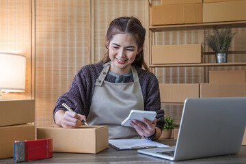 portrait of a young Asian woman, e-commerce employee sitting in the office full of packages in the background write note of orders and a calculator, for SME business ecommerce and delivery business.