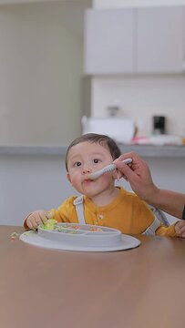 Boy having breakfast at the table at home while his grandmother feeds him with a spoon