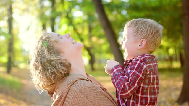 A little boy is having fun and fooling around with his mom. Close-up video portrait of a mother and son during a walk in the autumn park. Woman holds her toddler son in her arms and hugs him tenderly.