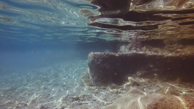 Camera moving forwards along large stones in coastal area on bright sunny day in sun glare, a school of mullet fish swims past. Underwater landscape of Mediterranean Sea
