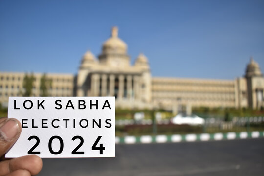 A closeup picture of a card with text- Lok Sabha elections 2024, held in front of parliament building.