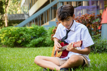 Asian schoolboy in school uniform playing ukulele or acoustic guitar lonely in school park, asian boy with popular intruments concept.