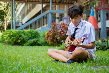 Asian schoolboy in school uniform playing ukulele or acoustic guitar lonely in school park, asian...