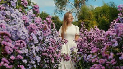 Caucasian woman with long hair in white dress walks among vibrant purple wildflowers in spring...