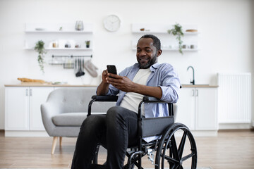 Close up view of african male with disability using smartphone while resting in modern kitchen of apartment. Joyful person in casual clothes texting message on digital device at home