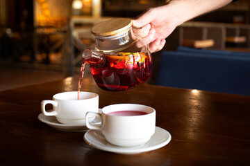 A man pours black fragrant fruit tea from a teapot into 2 cups. Tea made of orange, cinnamon,...