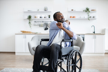 Joyous bearded man in wheelchair exercising stretching arms while following workout program at home. Positive african adult in cozy outfit participating in physical activity in apartment.