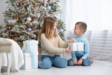 a boy and a girl give each other gifts against the background of a New Year tree. happy brother and sister opening New Year's gifts