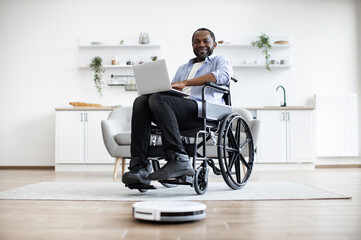Robotic vacuum removing dust from carpet while african homeowner with mobility impairment using laptop. Freelancer in wheelchair leading comfortable lifestyle by using tech gadgets