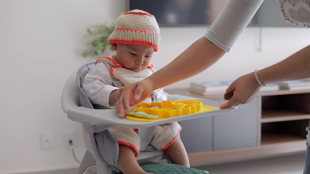 A baby sitting in his chair receives his plate of food for breakfast at home. Baby led weaning concept