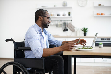 African bearded male in wheelchair using computer during mealtime in kitchen interior. Business...