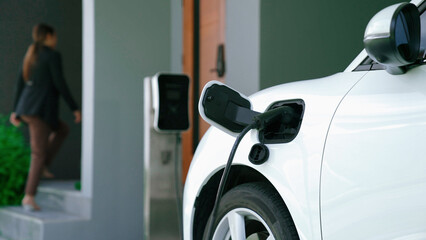 Progressive woman installs a charging station plug into her electric vehicle at home. EV...