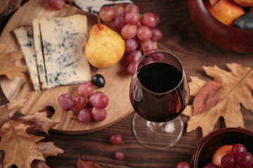 Glass of red wine served with blue cheese on dark wooden background. Autumn picnic with wine and cheese platter, fruits and dry leaves in rustic style. Top view