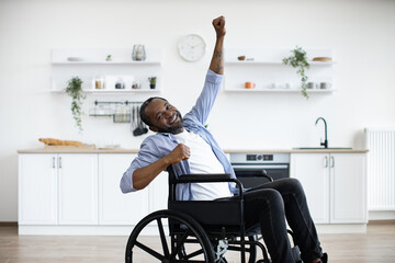 Full length portrait of african american wheelchair user smiling and dancing with raising hands while resting in kitchen of modern apartment. Young male in casual clothes spending pastime indoors. - obrazy, fototapety, plakaty