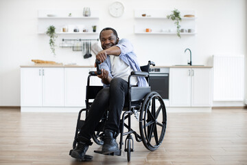 Full length portrait of african american wheelchair user with video game controller resting in open-plan kitchen of modern apartment. Cheerful young male in casual clothes spending pastime indoors.