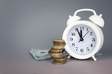 White alarmclock with Coins stack and stack of one hundred dollar bills on grey background. Deadline and time is money concept with White alarmclock and coin currency.