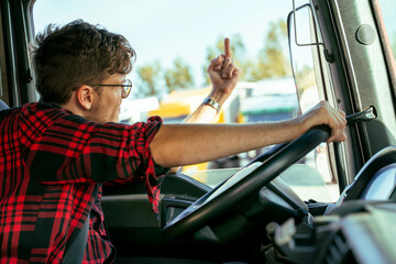Angry young truck driver showing middle finger to somebody closeup shot from cabin, road rage or job dissatisfaction  - obrazy, fototapety, plakaty