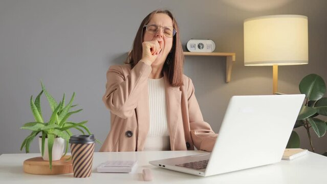 Computer work. Home office. Remote job. Notebook work. Sleepy tired woman using laptop working remotely at home office workplace yawning feeling sleepless drinking coffee to have energy.