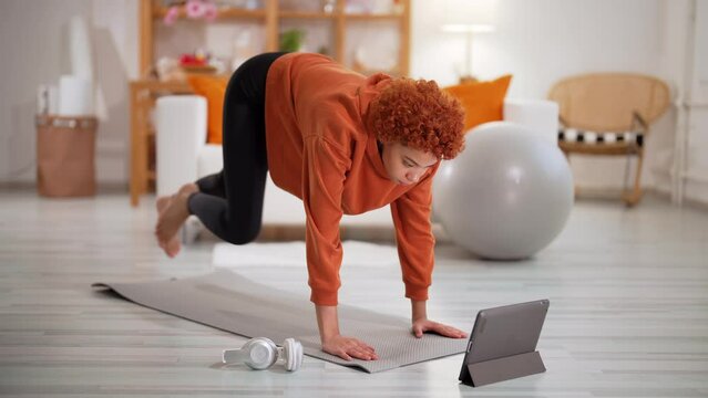 Fit slim african american young girl in sportswear doing legs exercise in plank pose at home on mat. Curly redhead woman training online using tablet. Workout, fitness, online exercising concept.