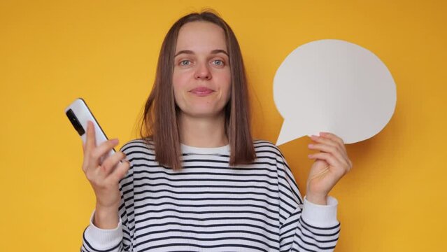 Puzzled woman wearing striped shirt posing with blank speech bubble for your promotional text against yellow wall holding smartphone shrugging shoulders doesn't know what comment to leave.