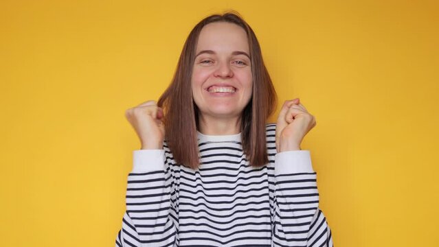 Extremely happy woman wearing striped casual shirt posing isolated over yellow background being overjoyed clenched fists rejoicing good news celebrating.