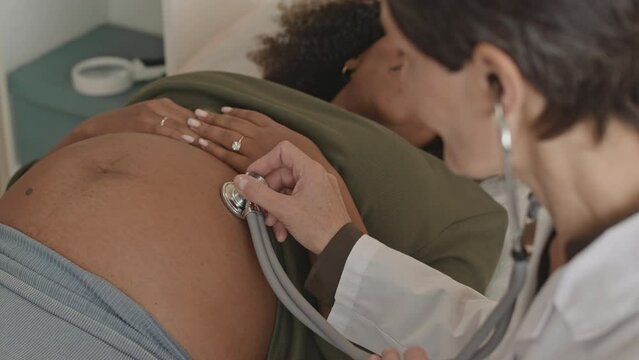 Medium closeup of young African American woman having her belly examined with stethoscope by mid adult Caucasian female doctor in modern clinic