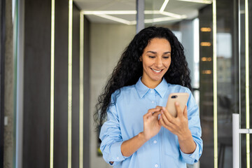 Young beautiful Inian woman stands in the office corridor of the hall, uses the phone, smiles,...