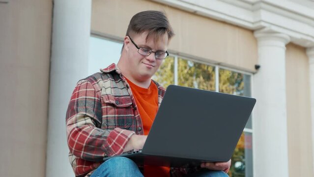 Young man with down syndrome in checkered shirt using laptop working online outdoors in the city. Guy with disability working remote online as freelancer