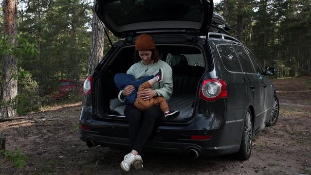 Young Woman Mother 30 Years Old In Hiking Clothes Sits On The Trunk Of A Car With A Child In Her Arms, Breastfeeds And Looks At The Sea, Road Trip, Tourism With A Child