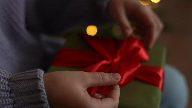 Close-up hands of unrecognizable little girl holding and using red ribbon to tie bow for wrapped gift box for Christmas present, on blurred background bright bokeh lights of Xmas tree, slow motion.