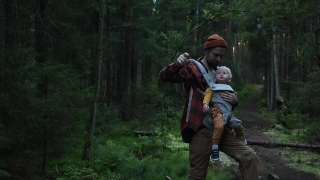 a male father with a baby in a sling-ergo backpack points his finger into the distance and points with his hand