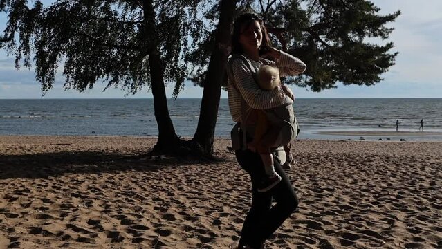 young woman mother walks along the beach against the sun by the sea, sun rays, shadows, wind, looks at the camera and smiles