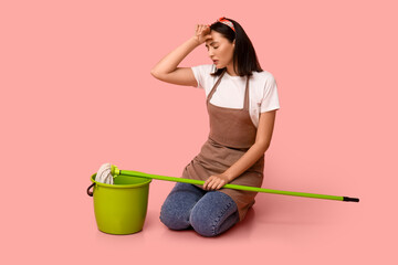 Portrait of tired young housewife in apron with mop and bucket on pink background - obrazy, fototapety, plakaty