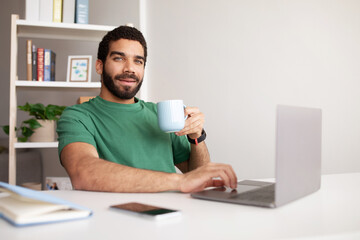 Positive millennial middle eastern guy work at computer, drink cup of tea, enjoy coffee break