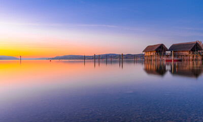 A serene sunrise scene by a tranquil lake with a small house on the shore. The sky is a beautiful...