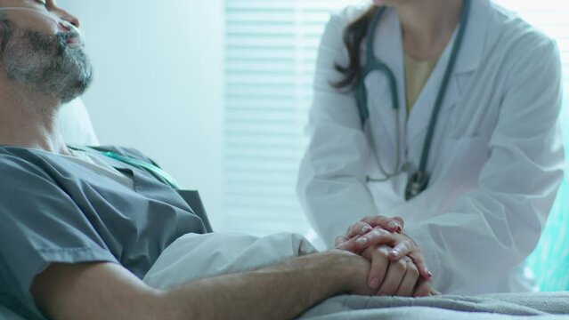 Young female doctor in white coat holding hands of male patient, smiling and talking with him, supporting and cheering up during treatment in hospital ward