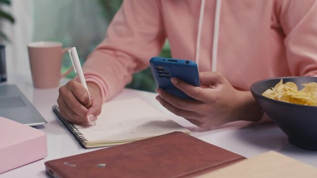 Medium shot of female teenager preparing for exam using smartphone and making notes while studying from home