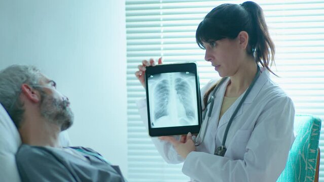 Young female doctor showing chest x-ray on digital tablet and zooming image while explaining diagnosis to male patient lying on bed in hospital ward
