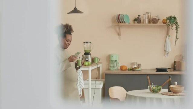 Medium shot of happy young Black pregnant woman taking pictures of herself drinking homemade green smoothie during lunchtime in cozy apartment with pastel peach walls