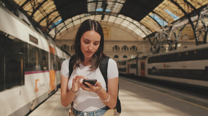 Beautiful girl traveler uses mobile phone while standing on the platform of the railway station