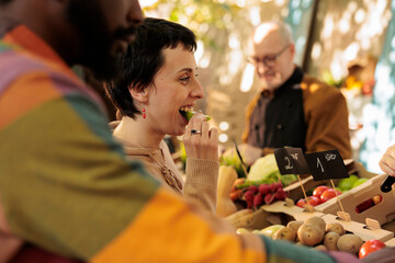 Vendor offering samples to customers while selling homegrown fruits and vegetables at local farmers market. Young multiracial family couple tasting natural organic produce while visiting food fair