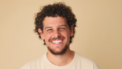 Close up, guy with curly hair dressed in beige t-shirt looks at the camera and laughs isolated on...